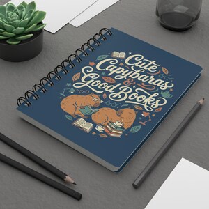 Può includere: Un quaderno a spirale blu navy con la scritta "Cute Capybaras & Good Books". Il design include illustrazioni di capibara, libri e altri elementi decorativi. Sono visibili anche una pianta succulenta e delle matite.