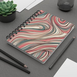 Può includere: Quaderno a spirale con un colorato design astratto. La copertina presenta linee fluenti nei toni del rosa, blu, beige e marrone. Il quaderno è aperto, rivelando pagine bianche. Sono visibili anche due matite e una pianta succulenta.