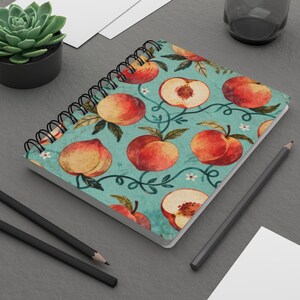Può includere: Quaderno a spirale con copertina turchese decorata con un motivo di pesche e tralci. Le pesche sono raffigurate intere e tagliate. Sullo sfondo ci sono matite nere e una piccola pianta succulenta.