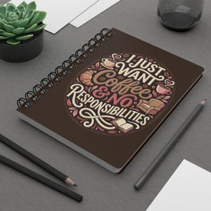 Può includere: Un taccuino marrone a spirale con il testo "I Just Want Coffee & No Responsibilities" in un design decorativo. Il taccuino è su una superficie grigia con matite e una piccola pianta in vaso.