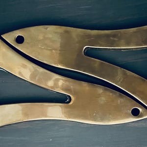 以下が含まれることがあります： 真鍮色の装飾用金属部品が2つ組み合わさり、対称的なデザインを形成しています。各部品は、切り抜かれたセクションと小さな隆起した足を持つ、湾曲した有機的な形状をしています。 全体的な美学はヴィンテージまたはアンティークです。