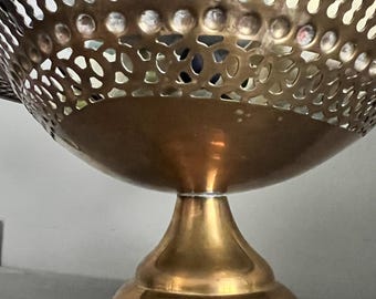 May include: A decorative brass bowl with a pedestal base. The bowl has a perforated design around the top and a smooth, rounded base. The brass has a warm, golden hue, and the bowl is likely used for display or serving.