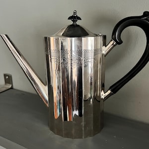 May include: A silver-colored coffee pot with a black handle and lid finial. The pot has a long spout and a decorative band around the middle. The handle is curved and the lid has a small, ornate knob.