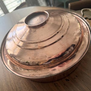 May include: A copper cooking pot with a lid. The pot has a round shape with a series of concentric rings on the lid. It features two brass handles on either side. The pot is sitting on a wooden surface.
