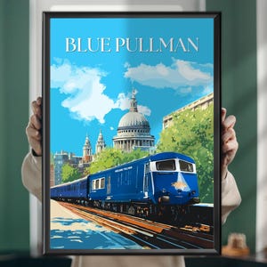 Op de afbeelding: Een ingelijste print met een blauwe "BLUE PULLMAN" trein tegen een achtergrond van een stadsgezicht, met de St Paul's Cathedral. Het kunstwerk heeft een vintage reisposterstijl, met een blauwe lucht en witte wolken.