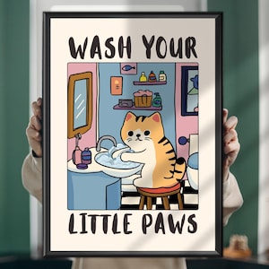 Può includere: Una stampa a fumetti incorniciata con un gatto che si lava le zampe in un lavandino del bagno. La stampa ha le parole "WASH YOUR LITTLE PAWS" in grassetto nero. Il gatto è arancione e bianco e il bagno ha pareti rosa e blu.