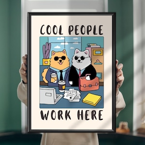 Op de afbeelding: Een ingelijste kunstprint met een cartoonillustratie van twee honden in pakken en zonnebrillen die aan een bureau zitten met koffie, een laptop en een aktetas. De tekst COOL PEOPLE WORK HERE staat in het zwart boven- en onderaan gedrukt.