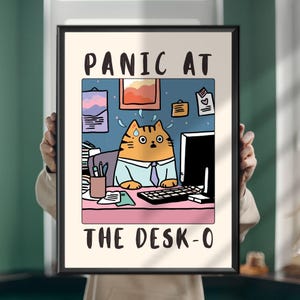 May include: A framed wall art print featuring a cartoon orange tabby cat wearing a light blue shirt and purple tie at a desk with a computer, keyboard, and office supplies. The text PANIC AT THE DESK-O is printed in black at the top and bottom.