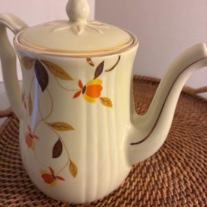 May include: A cream-colored ceramic teapot featuring a vertical ribbed texture and a floral pattern with orange, yellow, and brown leaves. The teapot includes a matching lid with a pointed finial and gold trim accents on the rim and spout.
