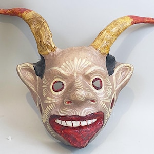May include: A handcrafted mask with a devilish design. The mask features curved horns with red tips, a smiling red mouth, and eye holes. The face is painted in shades of tan and white, with decorative patterns. The mask is likely used for costume or decorative purposes.