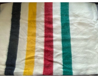 May include: A cream-colored wool blanket featuring a series of four vertical stripes in black, yellow, red, and green. The fabric has a soft, fuzzy texture throughout, with the colored stripes running parallel across the width of the textile.