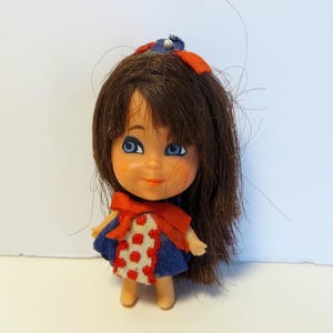 May include: A small doll with brown hair, blue eyes, and a smiling expression. The doll wears a blue dress with red polka dots and a red bow. A small blue and red bow adorns the doll's hair.