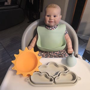 May include: A baby doll wearing a floral print dress and a green bib sits in a high chair. A bright orange sun-shaped plate, a gray cloud-shaped plate, and a light blue water bottle are on the tray.