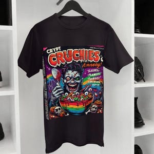 Könnte beinhalten: Schwarzes T-Shirt mit einer farbenfrohen Grafik eines Zombies, der einen Löffel über einer Schüssel mit Augäpfeln und Müsli hält. Die Grafik ist wie eine Müslipackung gestaltet, mit den Worten "Crypt Cruchies" und anderem Text.