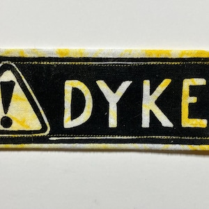 May include: A rectangular patch with a black background and yellow border. The word "DYKE" is in white capital letters. A yellow triangle with an exclamation point is on the left side of the patch.