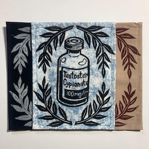 Peut inclure: Trois écussons en tissu rectangulaires présentant une illustration centrale à l'encre noire d'un flacon de médicament étiqueté "Testosterone Cypionate 100mg/ml" encadré de feuilles de laurier. Les tissus sont noir, bleu clair chiné et beige.