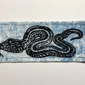 May include: A rectangular fabric patch featuring a black ink block print of a coiled snake with a forked tongue. The background fabric is a light blue and white mottled pattern, and the entire patch is finished with a dark blue stitched border.