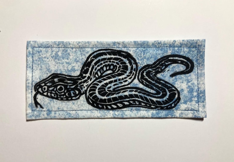 May include: A rectangular fabric patch featuring a black ink block print of a coiled snake with a forked tongue. The background fabric is a light blue and white mottled pattern, and the entire patch is finished with a dark blue stitched border.