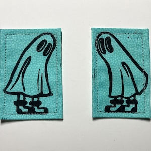 Könnte beinhalten: Zwei türkisfarbene Stoffaufnäher mit einem schwarzen Geisterdesign. Die Geister haben ovale Augen und tragen Stiefel. Die Aufnäher sind rechteckig mit einem gepunkteten Rand.