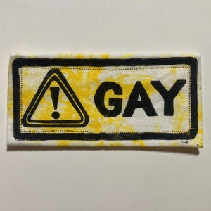 May include: A rectangular patch with a yellow and white background and black border. The patch features a black exclamation mark inside a yellow triangle, and the word "GAY" in bold black letters.