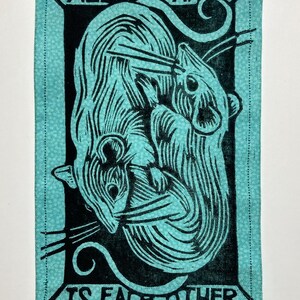 May include: A rectangular teal fabric patch with a black linocut print of two stylized rats intertwined. The text "ALL WE HAVE IS EACH OTHER" is printed in black around the image. The edges are stitched.