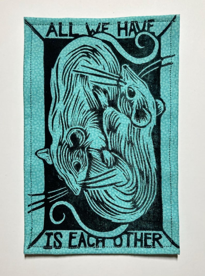May include: A rectangular teal fabric patch with a black linocut print of two stylized rats intertwined. The text "ALL WE HAVE IS EACH OTHER" is printed in black around the image. The edges are stitched.