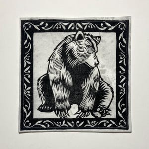Peut inclure: Estampe en linogravure noir et blanc d'un ours assis dans un cadre décoratif. L'ours est représenté avec des détails de fourrure et une expression sereine. Le cadre présente un motif floral orné et répétitif.