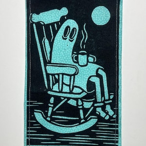 May include: A black and turquoise patch depicting a ghost in a rocking chair with a mug, under a full moon. The fabric patch has a turquoise border. The design is a linocut style.