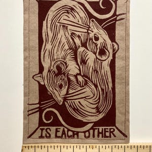 May include: A rectangular fabric patch with a brown illustration of two mice intertwined, with the text "ALL WE HAVE IS EACH OTHER" in a matching brown color. The patch is on a tan background.