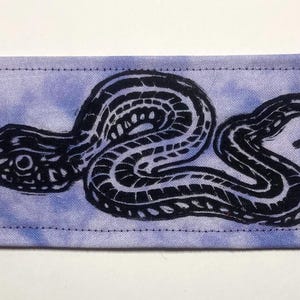 May include: A rectangular fabric patch featuring a tie-dye style light purple and white background with a black ink-style graphic print of a coiled snake. The patch has visible stitching along all four edges, creating a clean, finished border.