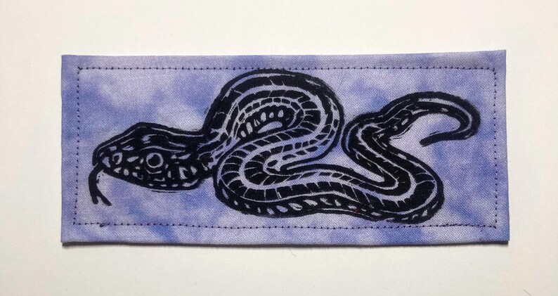 May include: A rectangular fabric patch featuring a tie-dye style light purple and white background with a black ink-style graphic print of a coiled snake. The patch has visible stitching along all four edges, creating a clean, finished border.