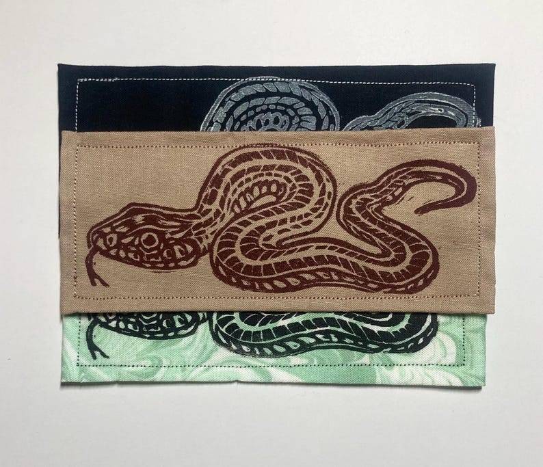 May include: Three rectangular fabric patches stacked vertically. The top patch is black with a grey snake print. The middle patch is tan with a dark brown snake print. The bottom patch is light green with a black snake print. Each has stitching.