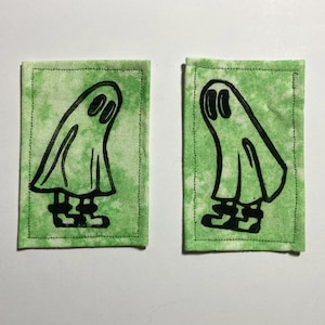 May include: Two rectangular patches featuring a green tie-dye pattern and a black ghost illustration. The ghost has oval eyes and blocky feet. The patches are outlined with black stitching.
