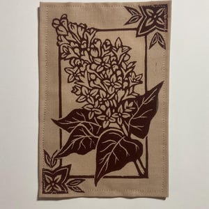 May include: A rectangular tan fabric piece featuring a dark brown block print of a lilac flower cluster with leaves. The design is enclosed within a thin rectangular border, with decorative floral corner accents and visible stitching along the edges.