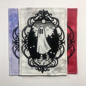 May include: Three rectangular fabric patches featuring a black screen-printed design of a ghost inside an ornate oval frame. The patches are layered, showing a purple tie-dye background, a white marble-effect background, and a red background.