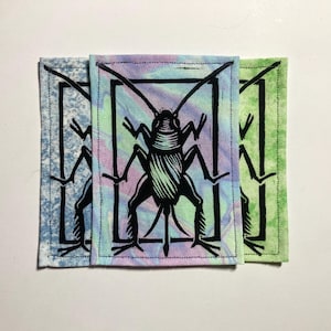 May include: Three fabric patches featuring a black insect design within a black rectangular frame. The patches have different backgrounds: blue, purple and green. The patches are rectangular with a stitched border.