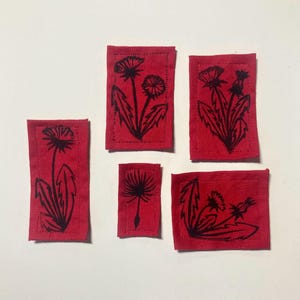 Op de afbeelding: Vijf rode stoffen patches met zwarte inkt illustraties van paardenbloemen. De patches zijn van verschillende groottes en vormen.