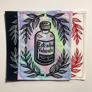 May include: Three fabric patches with botanical designs. The center patch features a black and white illustration of a medicine bottle with the text "Estradiol Valerate 40mg/m" surrounded by a laurel wreath. The other patches are black and white, and red and white.