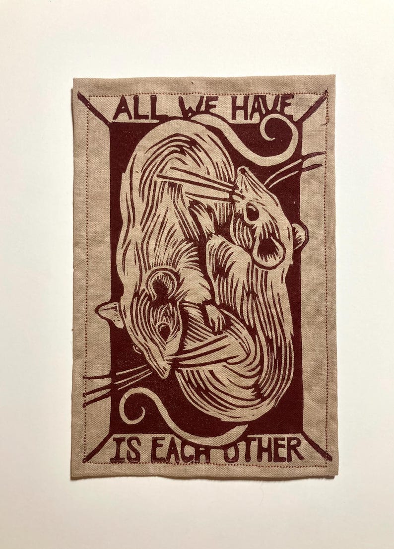 May include: A rectangular beige fabric patch with a maroon illustration of two mice intertwined. The text "ALL WE HAVE IS EACH OTHER" is printed in maroon around the image. The patch has a stitched border.