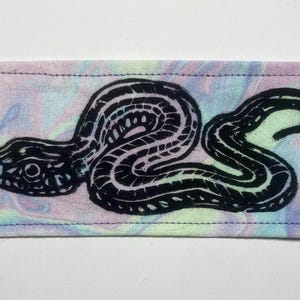 May include: A rectangular fabric patch featuring a black ink block print of a coiled snake with a forked tongue. The background fabric has a marbled, tie-dye pattern with soft pastel swirls of light blue, pink, yellow, and green throughout the surface.