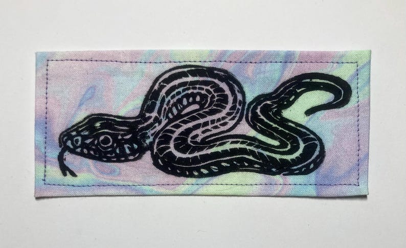 May include: A rectangular fabric patch featuring a black ink block print of a coiled snake with a forked tongue. The background fabric has a marbled, tie-dye pattern with soft pastel swirls of light blue, pink, yellow, and green throughout the surface.