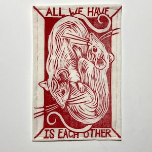 May include: A rectangular, off-white fabric art piece with a red linocut print of two stylized rats. The text "ALL WE HAVE IS EACH OTHER" is printed in red around the image. The piece has a dotted border.