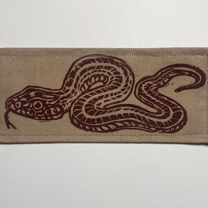 May include: A rectangular beige fabric patch featuring a dark brown block-printed graphic of a snake in a coiled position. The design shows the snake with a textured scale pattern and a forked tongue, with visible stitching along the edges.