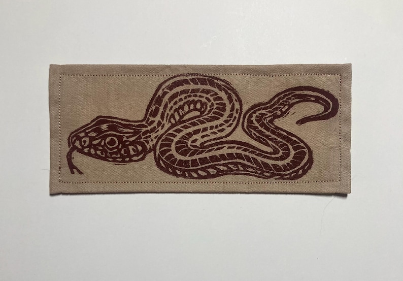 May include: A rectangular beige fabric patch featuring a dark brown block-printed graphic of a snake in a coiled position. The design shows the snake with a textured scale pattern and a forked tongue, with visible stitching along the edges.