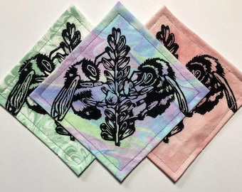 May include: Three square fabric coasters featuring a black ink block print of two bumblebees on a flower stalk. The left coaster is light green, the center is a pastel tie-dye blend, and the right coaster is a solid light pink fabric.