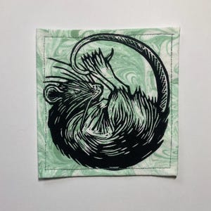 K&ouml;nnte beinhalten: Ein quadratischer Stoffuntersetzer mit einer schwarzen Linolschnitt-Illustration einer zusammengerollten Ratte. Die Ratte ist mit detaillierten Fellstrukturen und einem langen Schwanz dargestellt. Der Hintergrundstoff ist helles Minzgr&uuml;n.