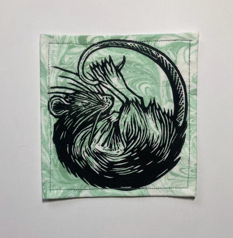 K&ouml;nnte beinhalten: Ein quadratischer Stoffuntersetzer mit einer schwarzen Linolschnitt-Illustration einer zusammengerollten Ratte. Die Ratte ist mit detaillierten Fellstrukturen und einem langen Schwanz dargestellt. Der Hintergrundstoff ist helles Minzgr&uuml;n.