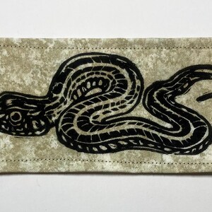 May include: A rectangular fabric patch featuring a black ink block print of a coiled snake with a textured, mottled beige background. The patch has visible stitching along all four edges, creating a clean border around the central serpent design.