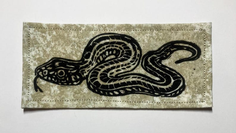 May include: A rectangular fabric patch featuring a black ink block print of a coiled snake with a textured, mottled beige background. The patch has visible stitching along all four edges, creating a clean border around the central serpent design.
