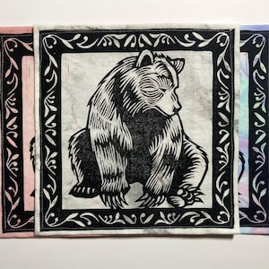 May include: Three square art prints with a black and white bear illustration in a decorative black frame. The prints have borders in pink, white, and blue. The bear is shown seated. Each print is approximately 20 cm x 20 cm.